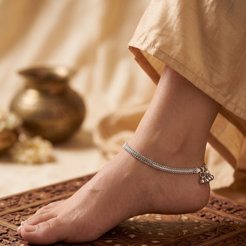 A silver chain anklet with a small hanging charm worn on a person's ankle, shown in a warm, styled setting with a beige garment and a carved wooden surface.