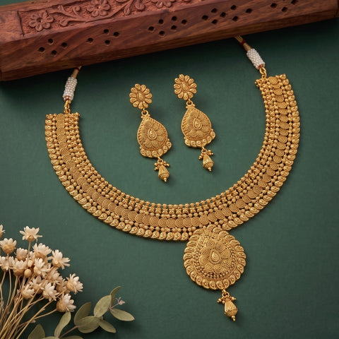 An ornate gold-toned traditional necklace and matching earrings displayed on a dark green background, with decorative dried flowers and a carved wooden prop at the top.