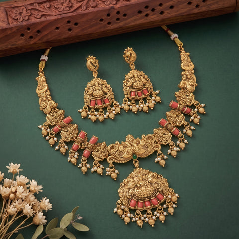 An ornate gold-toned traditional jewelry set with a necklace, earrings, and matching pendant, decorated with red stones and small pearl-like beads, arranged on a dark green background with a carved wooden prop and small white flowers.