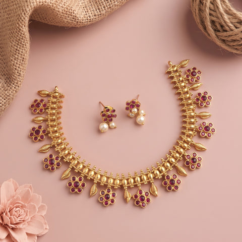Gold-toned traditional necklace and matching earrings set with red floral accents and pearl-like drops, arranged on a soft pink background with burlap and a pink flower prop.