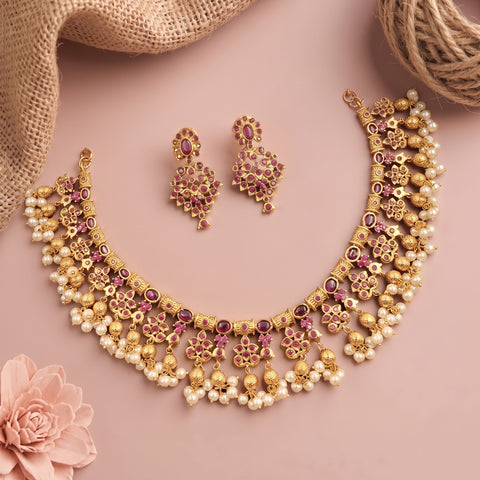 Gold-toned traditional necklace and matching earrings with pink gemstone accents and white pearl-like beads, arranged on a dusty pink background with burlap and a flower prop.