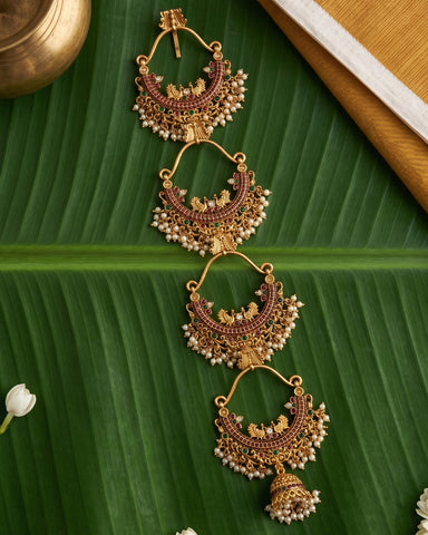 Four pairs of gold-toned traditional Indian jhumka earrings with red stone embellishments and white bead fringe, arranged on a green banana leaf with decorative props.