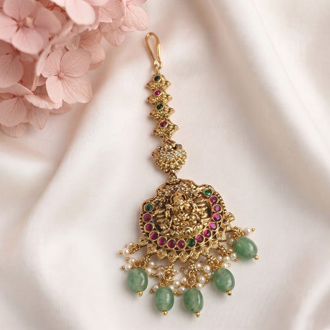 An ornate gold-toned traditional Indian choker necklace with a large Ganesha pendant, decorated with green stone beads, pink gemstones, and white faux pearls, displayed on a white satin fabric next to pink flowers.