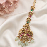 An ornate gold-toned traditional Indian jhumka earring decorated with pink and green stones, white pearl beads, and light green jade-like drops, displayed on a white satin fabric with pink flowers in the corner.