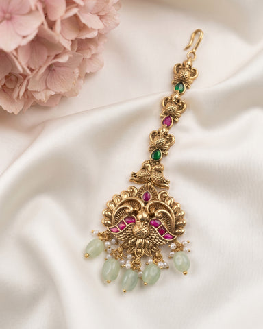 An ornate gold-toned traditional Indian jhumka earring decorated with pink and green stones, white pearl beads, and light green jade-like drops, displayed on a white satin fabric with pink flowers in the corner.