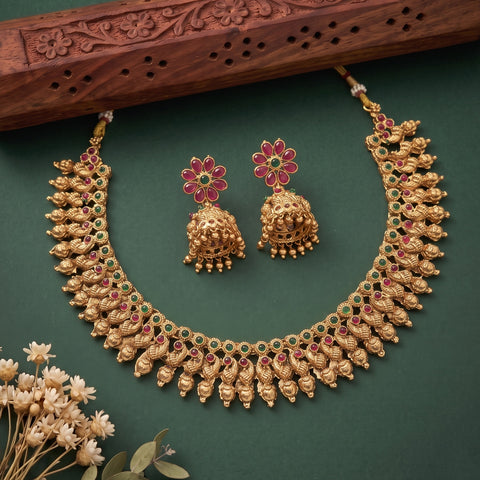 Gold-toned Indian jewelry set featuring a choker necklace and matching jhumka earrings, decorated with red and green stones, displayed on a dark green background with a carved wooden prop and small dried flowers.