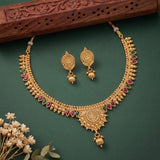 An ornate gold-toned traditional necklace and matching drop earrings with red and green stone accents, arranged on a dark green background with a carved wooden prop and small dried flowers.