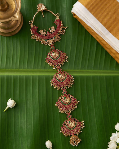 An ornate gold-toned traditional Indian necklace with red and green stone embellishments, displayed on a large green banana leaf with decorative props.