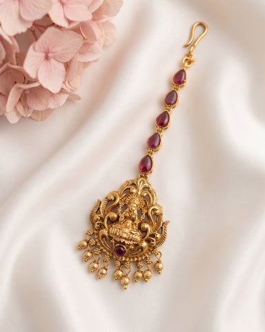 Gold-toned traditional Indian jewelry piece with a detailed Ganesha pendant, accented with deep red gemstones, displayed on a white satin fabric with pink flowers in the corner.