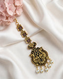 An ornate gold-toned traditional Indian jhumka earring with black enamel detailing, small purple gemstones, and dangling white faux pearls, displayed on draped white satin fabric next to pink flowers.