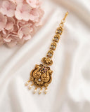 An ornate gold-toned traditional Indian jewelry piece, likely a maang tikka or hair pendant, decorated with small stones and hanging pearl-like drops, displayed on a white satin fabric next to pink flowers.