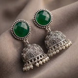 A pair of traditional Indian silver jhumka earrings with round green stone studs and dangling pearl-beaded bell-shaped charms, displayed on a draped taupe fabric background.