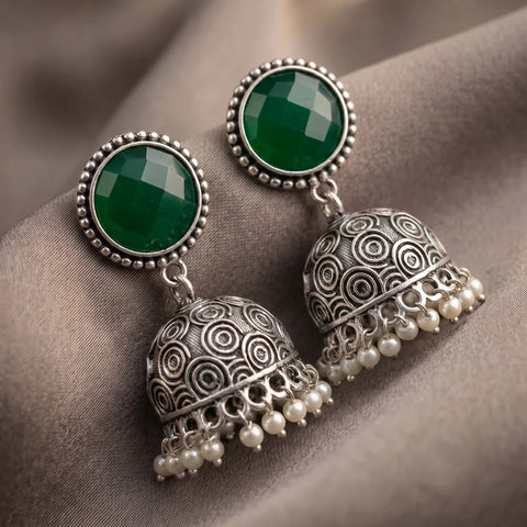 A pair of traditional Indian silver jhumka earrings with round green stone studs and dangling pearl-beaded bell-shaped charms, displayed on a draped taupe fabric background.
