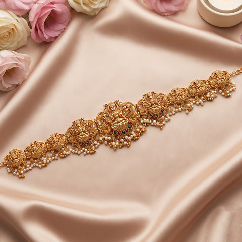 An ornate gold-toned traditional Indian bridal necklace with intricate filigree, red and green stone accents, and rows of white pearls, displayed on draped champagne satin fabric with decorative pink and cream roses.