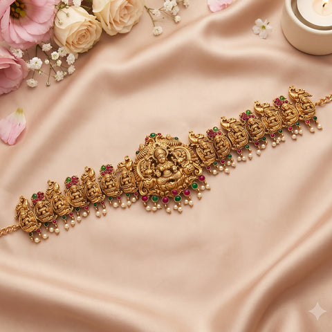 An ornate gold-toned traditional Indian choker necklace with detailed deity motifs, red and green stone accents, and dangling pearl beads, displayed on draped blush satin fabric with flowers and a candle in the corner.