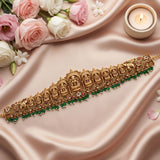 An ornate gold-toned traditional Indian choker necklace with detailed deity motifs, set against draped blush pink satin fabric, decorated with pink and white roses, a lit candle, and green bead accents.