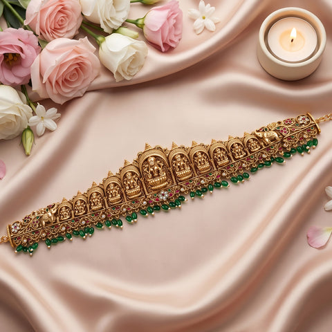 An ornate gold-toned traditional Indian choker necklace with detailed deity motifs, set against draped blush pink satin fabric, decorated with pink and white roses, a lit candle, and green bead accents.