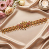 An ornate gold-toned traditional Indian choker or waist chain decorated with pink and green stones and dangling pearl beads, displayed on draped champagne satin fabric with roses and a lit candle in the corners.