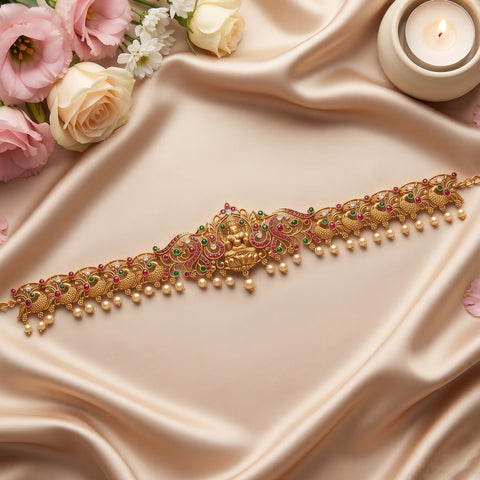 An ornate gold-toned traditional Indian choker or waist chain decorated with pink and green stones and dangling pearl beads, displayed on draped champagne satin fabric with roses and a lit candle in the corners.