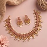 Gold-toned traditional jewelry set with a necklace, matching earrings, and pearl-like bead accents, arranged on a soft pink background with burlap and a pink flower prop.