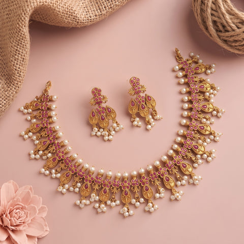 Gold-toned traditional jewelry set with a necklace, matching earrings, and pearl-like bead accents, arranged on a soft pink background with burlap and a pink flower prop.