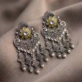 A pair of ornate silver-toned Indian-style dangle earrings with yellow enamel accents, clear stones, and small pearl-like beads, displayed on draped taupe fabric.