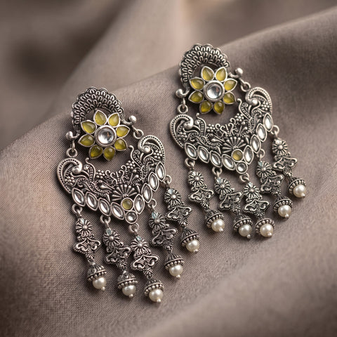 A pair of ornate silver-toned Indian-style dangle earrings with yellow enamel accents, clear stones, and small pearl-like beads, displayed on draped taupe fabric.
