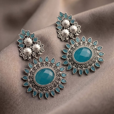 A pair of ornate silver-toned dangle earrings with large oval turquoise-blue center stones and smaller matching blue teardrop stones, displayed on draped taupe fabric.