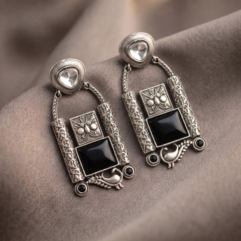 A pair of ornate silver-toned dangle earrings with black rectangular stones and clear heart-shaped studs, displayed on draped taupe fabric.