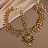 An ornate gold-toned traditional necklace set with matching pendant, displayed on a beige satin fabric background with small white flowers. The jewelry features red and green stone accents and a large decorative pendant.