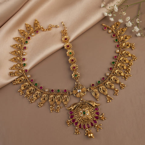 An ornate gold-toned traditional necklace set with matching pendant, displayed on a beige satin fabric background with small white flowers. The jewelry features red and green stone accents and a large decorative pendant.