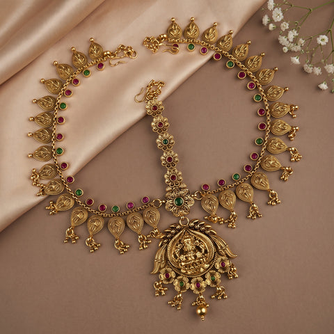 Gold-toned traditional Indian jewelry set featuring a necklace with a large deity pendant, a matching bracelet, and a collar, decorated with red and green stone accents, displayed on beige satin fabric.