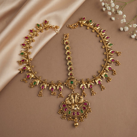 Gold-toned traditional Indian jewelry set featuring a necklace, matching bracelet, and a decorative pendant with red and green stone accents, arranged on a beige surface with satin fabric and small white flowers.