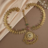 An ornate gold-toned traditional necklace with leaf-shaped chain details, a large circular pendant, and small red and green stone accents, arranged on draped beige satin fabric with white flowers in the corner.