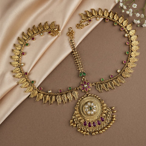 An ornate gold-toned traditional necklace with leaf-shaped chain details, a large circular pendant, and small red and green stone accents, arranged on draped beige satin fabric with white flowers in the corner.