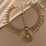 An ornate gold-toned traditional necklace with matching bracelet laid flat on a beige satin cloth. The jewelry features intricate filigree, hanging decorative elements, and multiple small red gemstone accents.