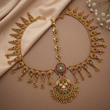 An ornate gold-toned traditional necklace with a large pendant, decorated with red and green stone accents, displayed on a beige satin fabric background with small white flowers in the corner.