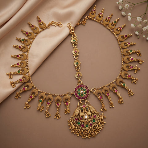 An ornate gold-toned traditional necklace with a large pendant, decorated with red and green stone accents, displayed on a beige satin fabric background with small white flowers in the corner.