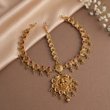 Gold-toned traditional Indian jewelry set including a necklace and matching earrings, decorated with red and green stone accents, displayed on a beige satin fabric background with small white flowers in the corner.