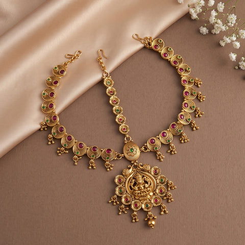 Gold-toned traditional Indian jewelry set including a necklace and matching earrings, decorated with red and green stone accents, displayed on a beige satin fabric background with small white flowers in the corner.
