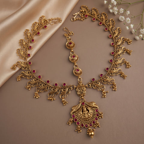 An ornate gold-toned traditional necklace set with matching earrings and a maang tikka, decorated with small red gemstone accents, displayed on a beige satin fabric with white baby's breath flowers in the corner.