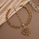 An ornate gold-toned Indian-style necklace with a large pendant and matching chain, decorated with red and green stone accents and small pearl-like details, arranged on a beige satin fabric backdrop with white baby's breath flowers in the corner.