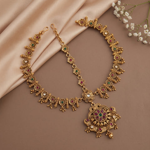 An ornate gold-toned Indian-style necklace with a large pendant and matching chain, decorated with red and green stone accents and small pearl-like details, arranged on a beige satin fabric backdrop with white baby's breath flowers in the corner.