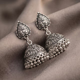 A pair of ornate silver-toned jhumka earrings with intricate floral engravings and dangling bead chains, displayed on draped taupe fabric.