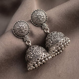 A pair of ornate silver-toned jhumka earrings with detailed filigree and beadwork, displayed on draped taupe fabric.