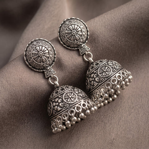 A pair of ornate silver-toned jhumka earrings with detailed filigree and beadwork, displayed on draped taupe fabric.