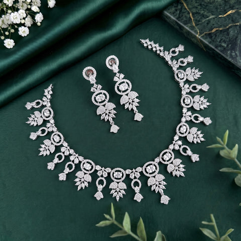 A silver-toned jewelry set featuring a necklace, matching earrings, and a small pendant, all decorated with intricate floral and circular motifs. The jewelry is displayed on a dark green satin fabric background with decorative green leaves and small white flowers.