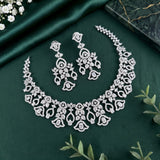A silver-toned jewelry set featuring a floral lace necklace and matching drop earrings, arranged on a dark green satin fabric background with decorative green leaves and small white flowers.