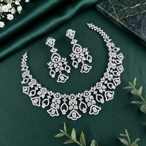A silver-toned jewelry set featuring a floral lace necklace and matching drop earrings, arranged on a dark green satin fabric background with decorative green leaves and small white flowers.