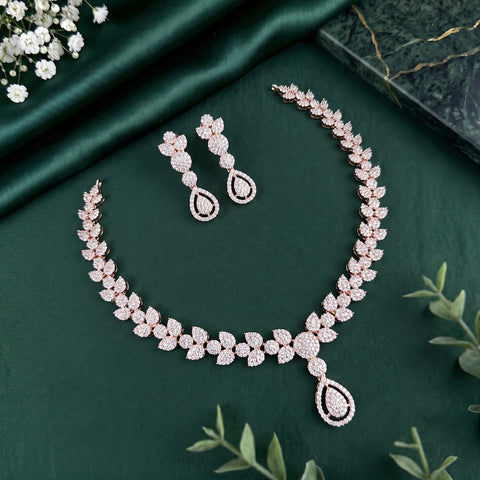 A rose gold jewelry set featuring a necklace and matching earrings, decorated with sparkling clear stones in a leaf and teardrop motif, displayed on a dark green satin fabric background.